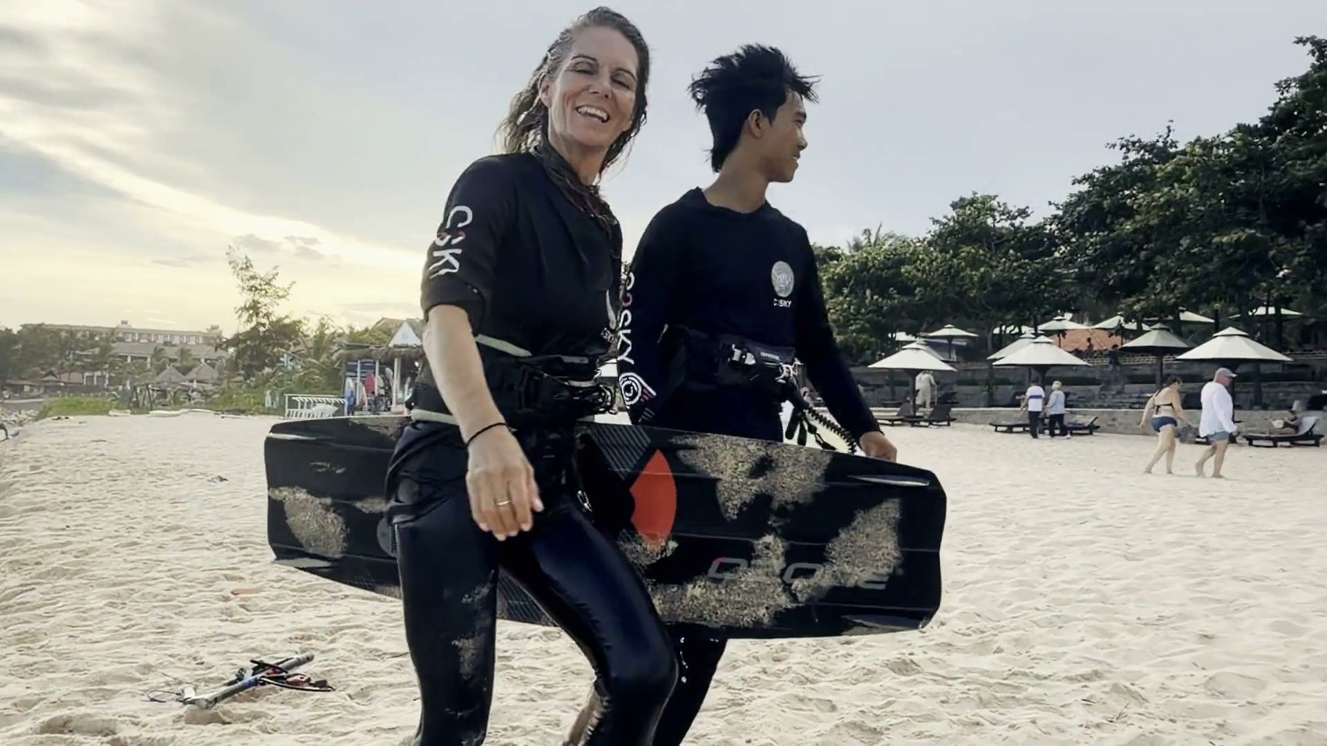 Liz and a C2SKY instructor laughing as they carry boards up the beach — play video