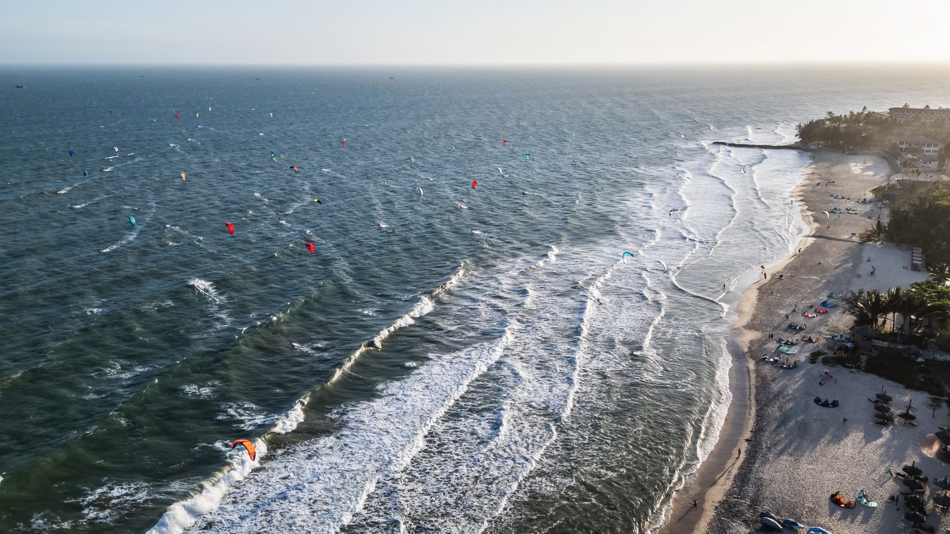 An aerial view along the bay at Mui Ne with dozens of kites out on the water and lines of surf running in to the beach.
