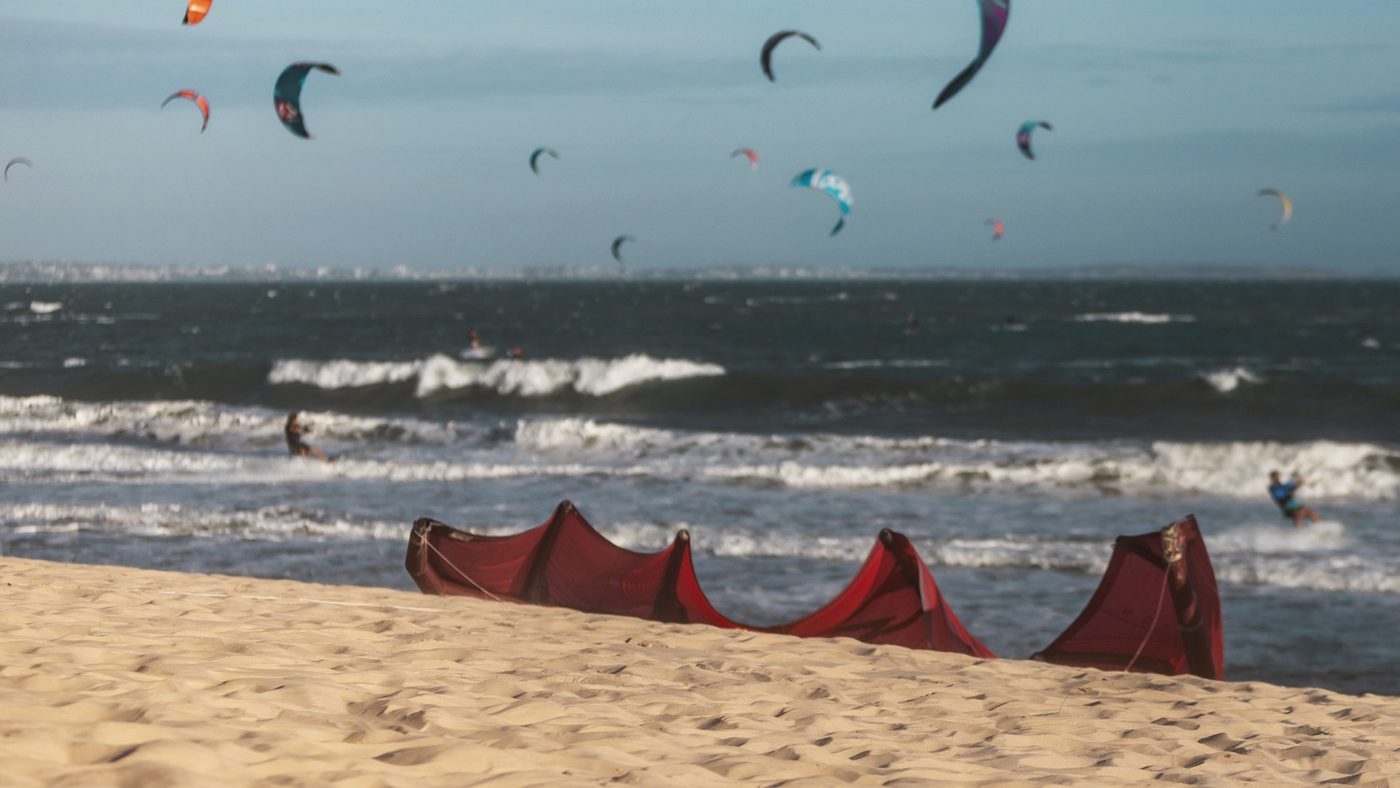 Kites rigged out along the sand, with more kites flying over the bay behind.