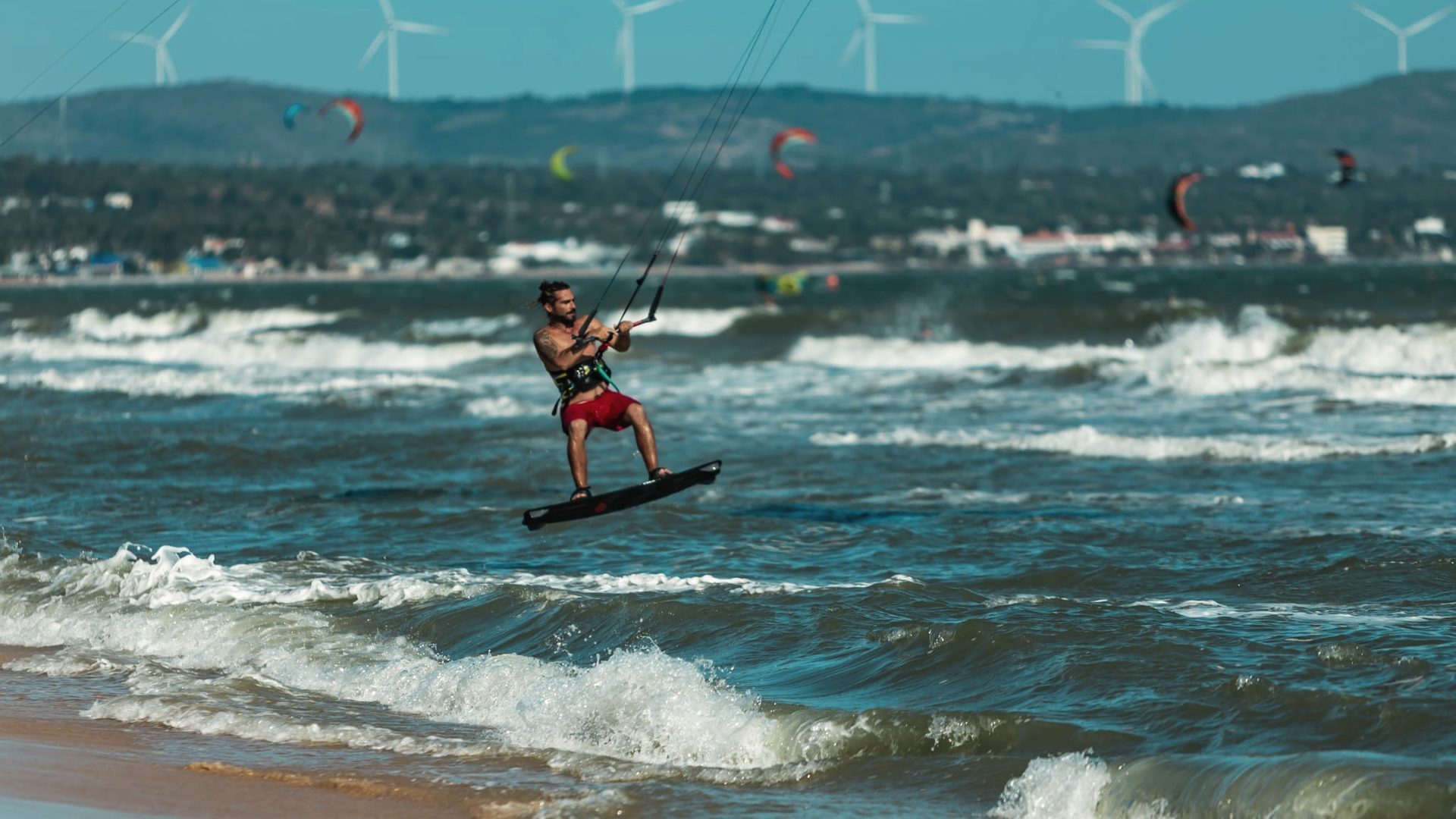 Ein Kitesurfer fährt durch die Brandung bei Mui Ne, dahinter die Windräder auf dem Bergrücken.