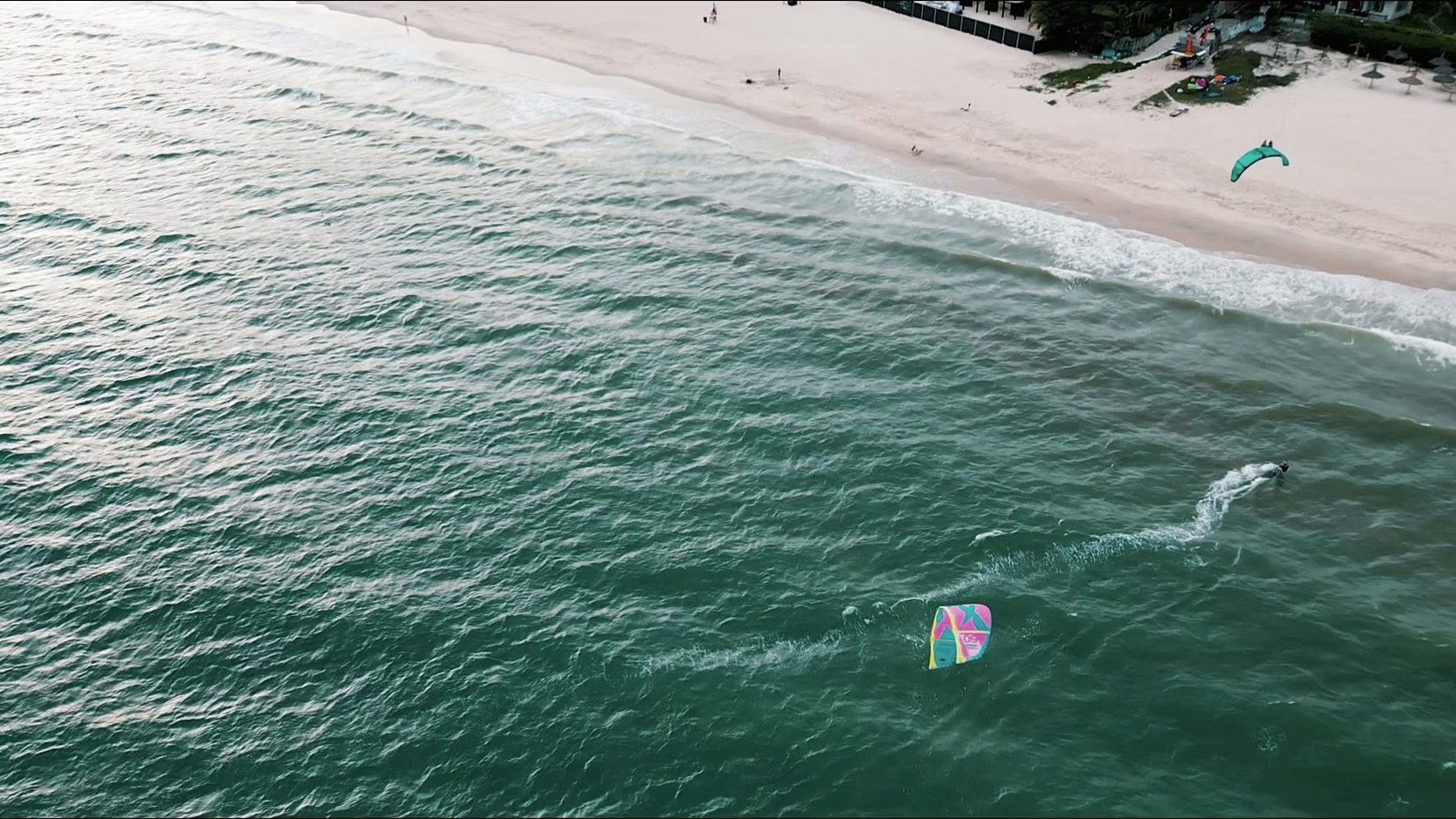 An aerial down the Mui Ne beach, a kitesurfer cutting a white wake across green water.