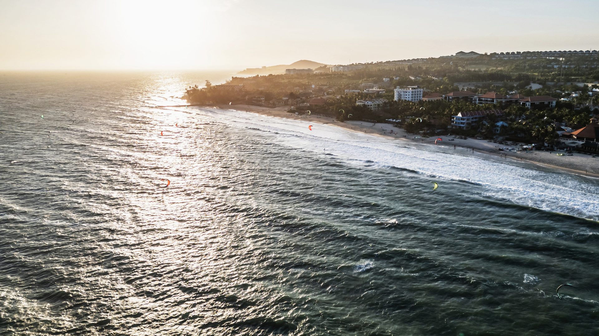 A golden-hour aerial down the Mui Ne coastline, kites out on the water and the resort strip in the distance.
