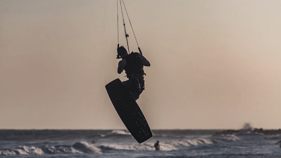 A kitesurfer in silhouette mid-jump above choppy water at dusk, board raised, kite arcing overhead against a soft peach sky.