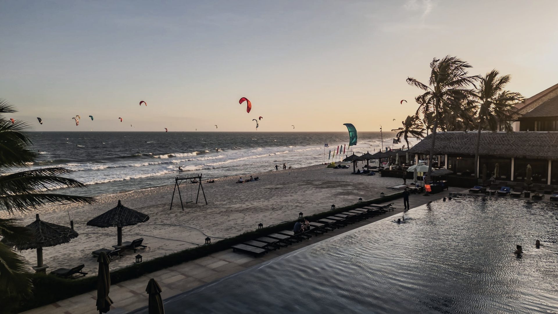 Sunset from a beachfront deck at Mui Ne, kites still in the air over the water, palms and parasols along the sand.