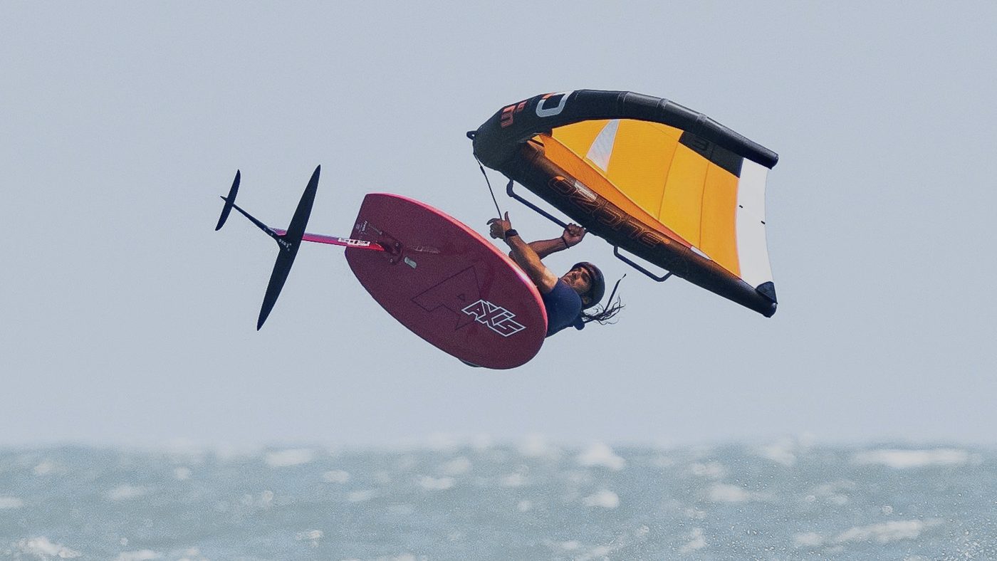 A wingfoiler clear of the water, board and foil lifted, orange wing held overhead off Mui Ne.