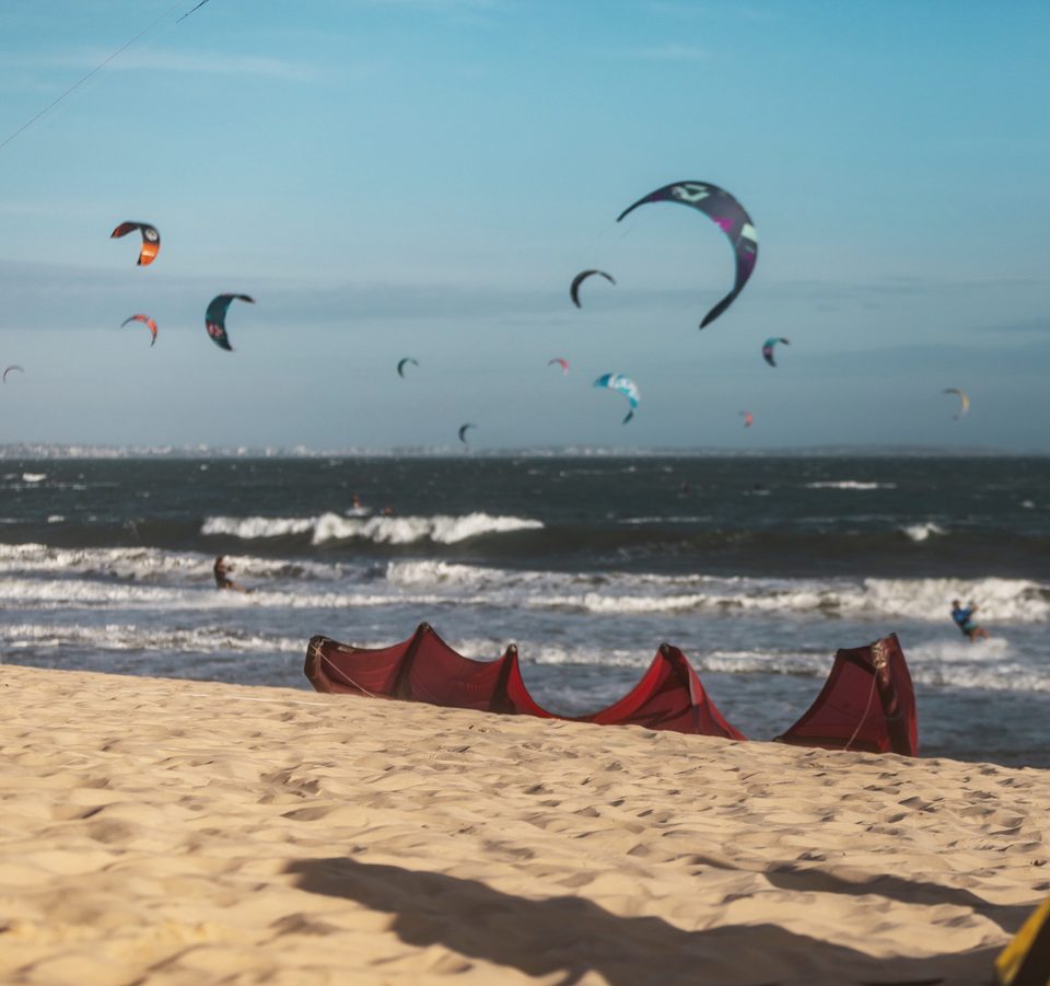 Kites rigged and laid out along the sand at C2SKY with their lines run out, and more kites flying over the water beyond.