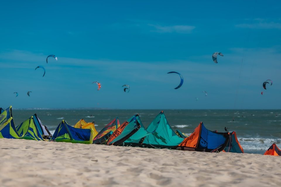 Kites laid out in a line along the sand at C2SKY, with a dozen more flying over the bay behind them under a clear sky.