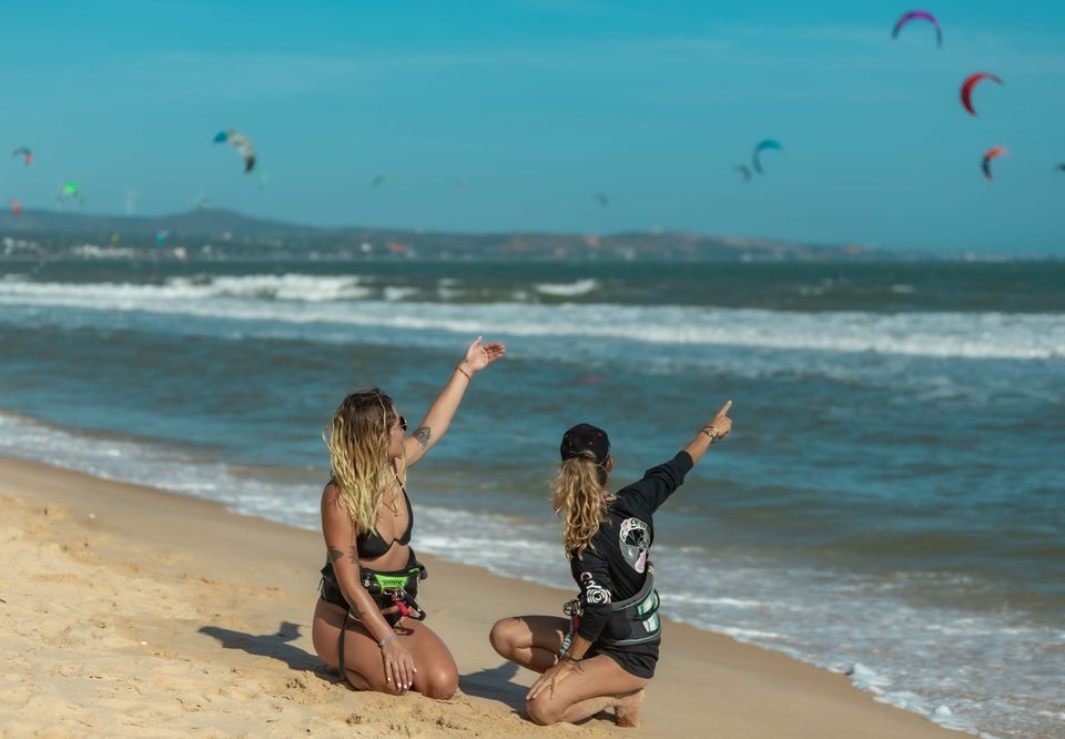 Two people kneeling on the sand at Mui Ne, one pointing up at the kites flying over the water, reading the conditions before rigging.