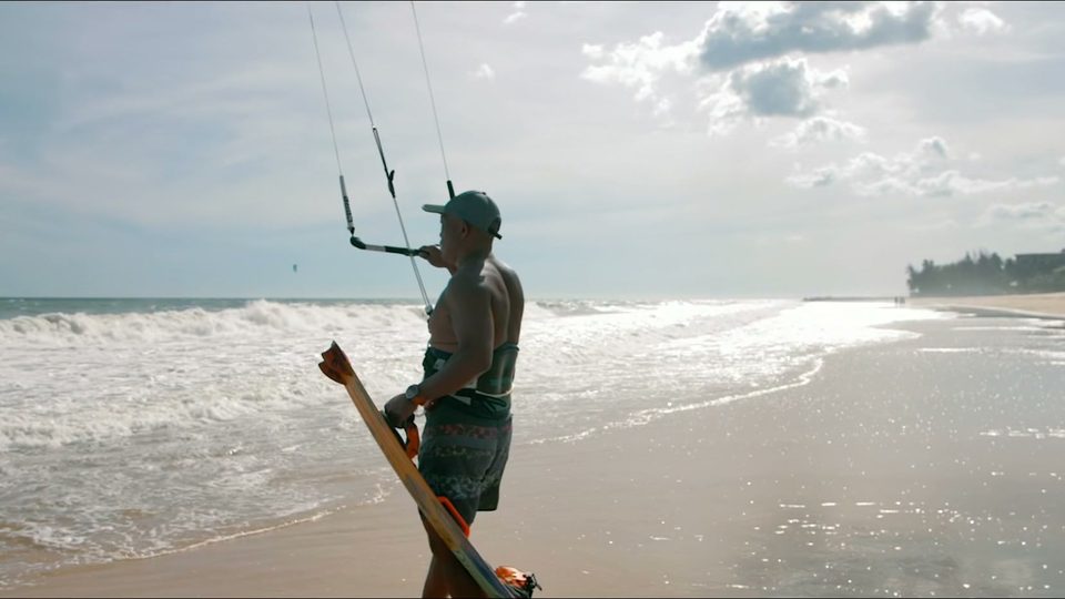A rider walking out along wet sand at Mui Ne carrying a board under one arm, kite lines running up out of frame, an empty beach stretching away behind.