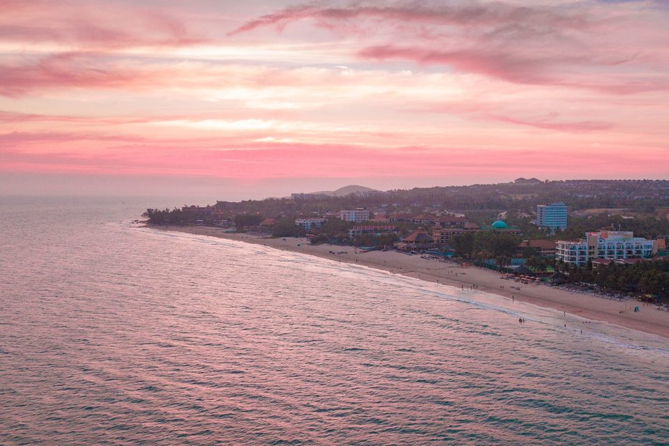 A sunset aerial along Mui Ne bay: a pink and lilac sky over a long curving beach, with the resort strip and palms running away into the haze.