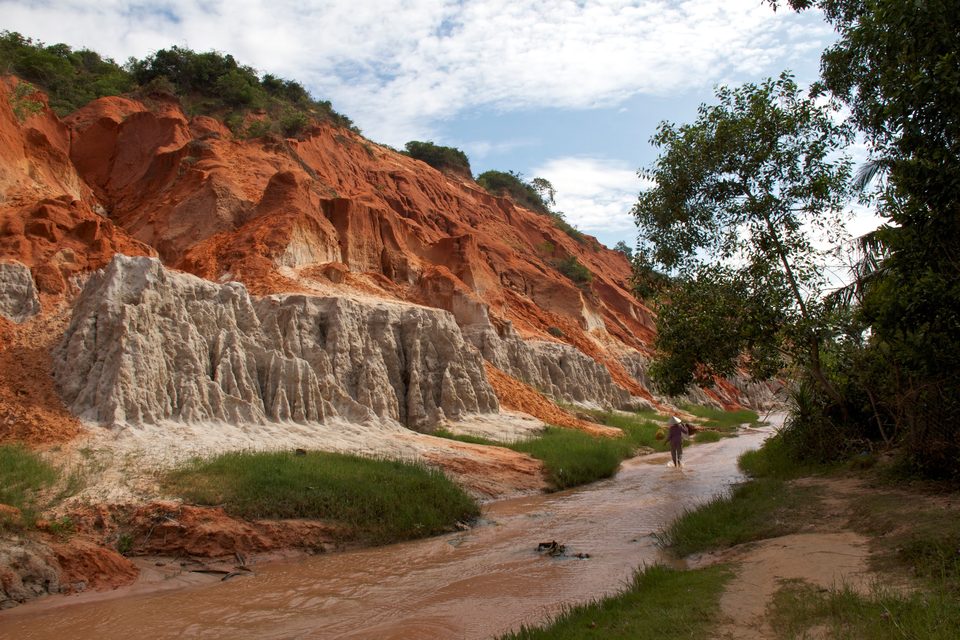 Fairy Stream at Mui Ne: a shallow stream running over pale sand between eroded red and white sandstone cliffs, with a person walking up it barefoot.