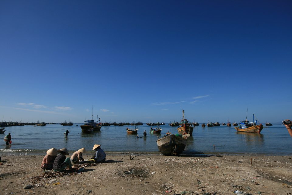 The fishing harbour at Mui Ne: a fleet of blue wooden boats at anchor in shallow water, round woven basket boats pulled up on the sand, and women in conical hats working the morning catch on the shore.
