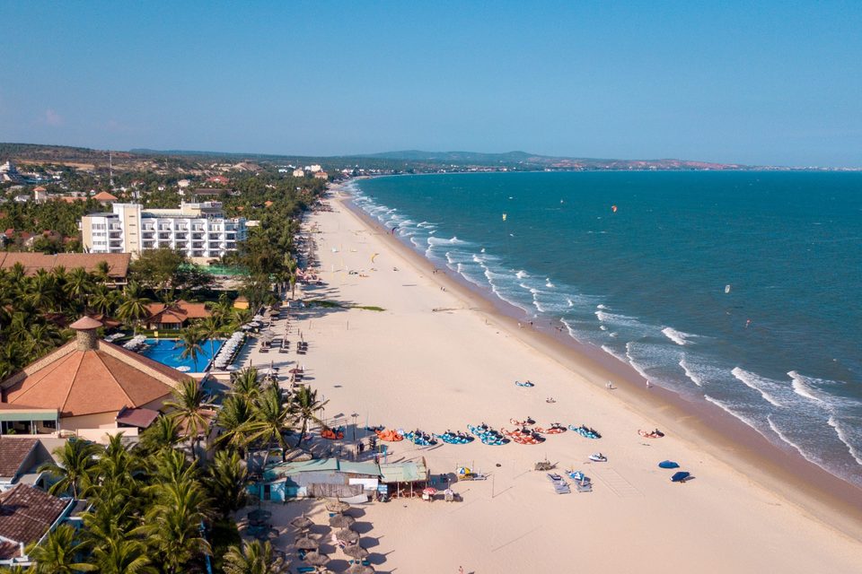 An aerial view along the Mui Ne strip: a line of resorts one building deep, a long pale beach, and kites out on the water beyond the surf.