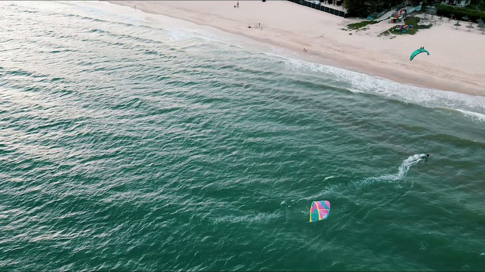 An aerial looking down the Mui Ne beach, a kitesurfer cutting a white wake across green water, one kite low over the surface and the resort strip along the sand.