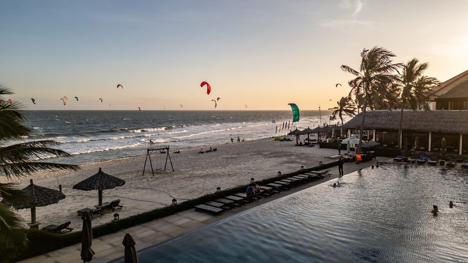 Sunset from a beachfront deck in Mui Ne: parasols and palms in silhouette, people on the sand, and kites still flying over the water.