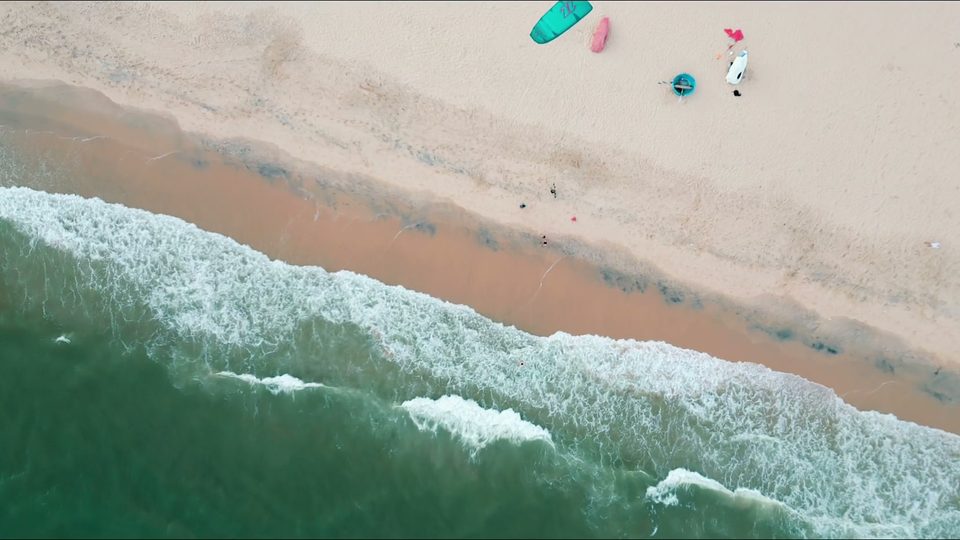 A straight-down aerial of the kite beach at Mui Ne: surf breaking onto pale sand, with kites laid out along the beach.