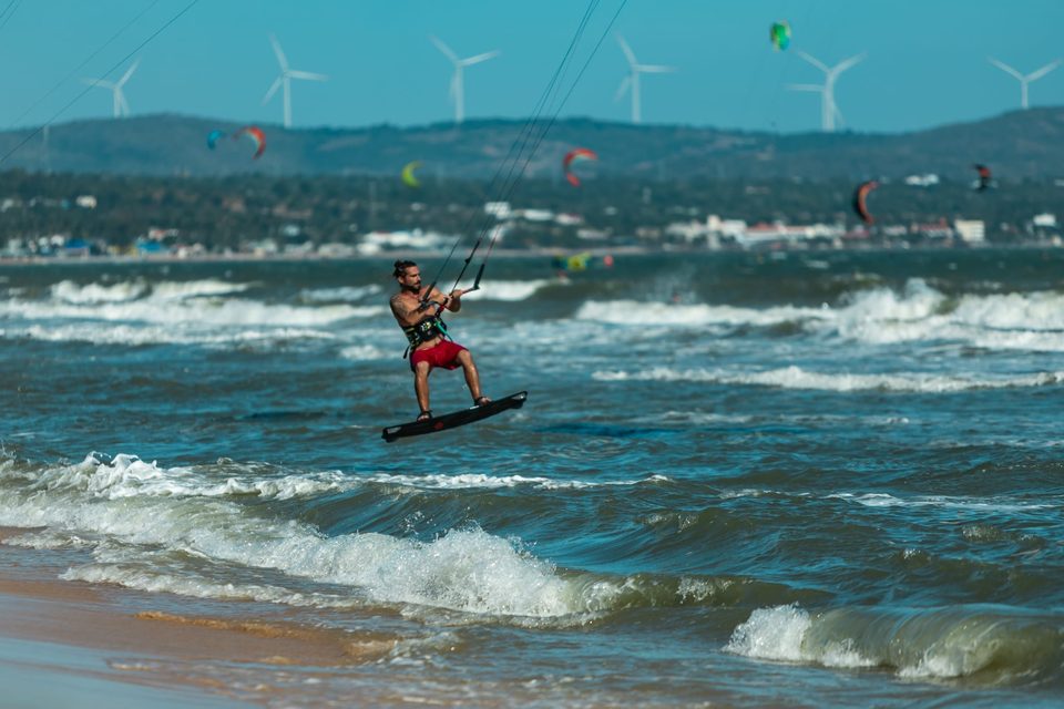 A kitesurfer riding across chop at Mui Ne, with a line of wind turbines standing on the ridge behind the bay.