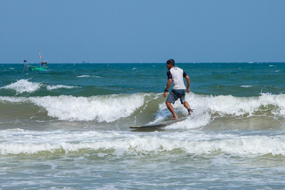 A rider on a small breaking wave at Mui Ne, with a fishing boat out on the flat water behind and a green headland in the distance.