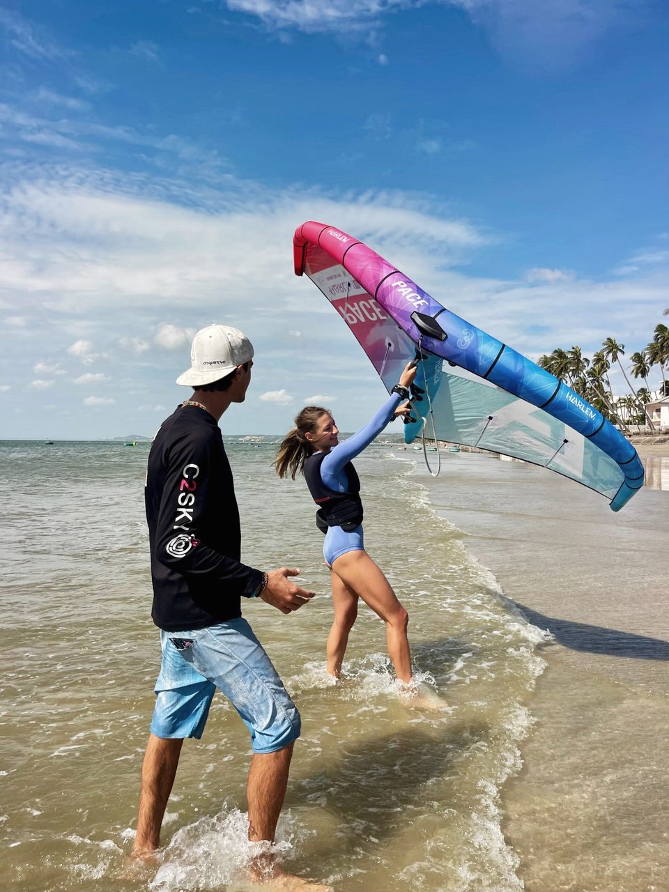 A C2SKY instructor coaching a student who is holding a wing overhead in shallow warm water at Mui Ne, palms along the shore behind.